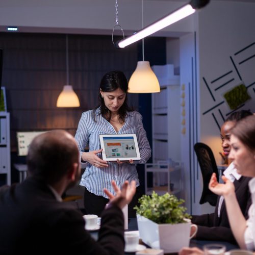 Overworked workaholic entrepreneur woman shwoing marketing graphs using tablet overworking at company solution late at night in meeting room. Diverse multi-ethnic teamwork solving financial strategy